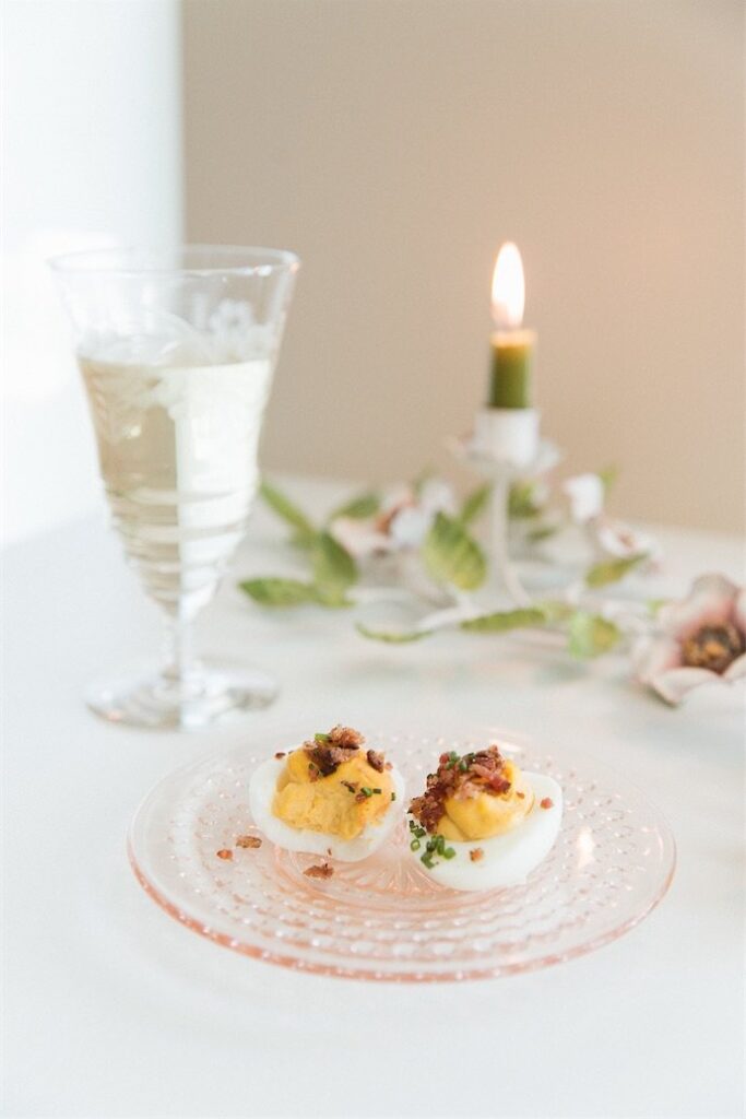 A glass of white beverage, a lit candle, and two deviled eggs garnished with herbs and bacon bits on a pink glass plate.