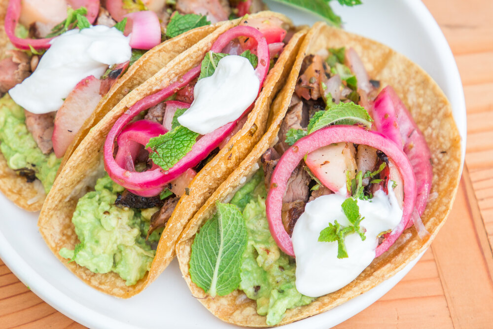 Three tacos on a white plate, filled with guacamole, grilled meat, pickled red onions, fresh mint leaves, and a dollop of sour cream.