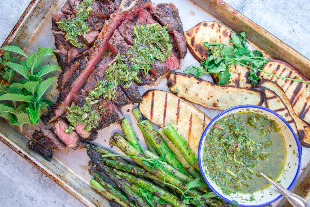 Grilled steak with green herb sauce, grilled eggplant, asparagus, a bowl of sauce with a spoon, and fresh greens arranged on a tray.