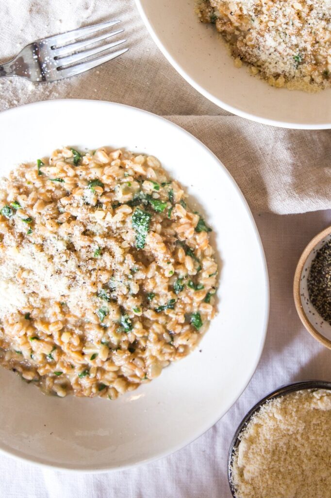 A white plate with a serving of creamy One-Pot Parmesan and Spinach Farrotto, topped with grated cheese and herbs, sits next to a fork, a bowl of more farrotto, grated cheese, and ground pepper.