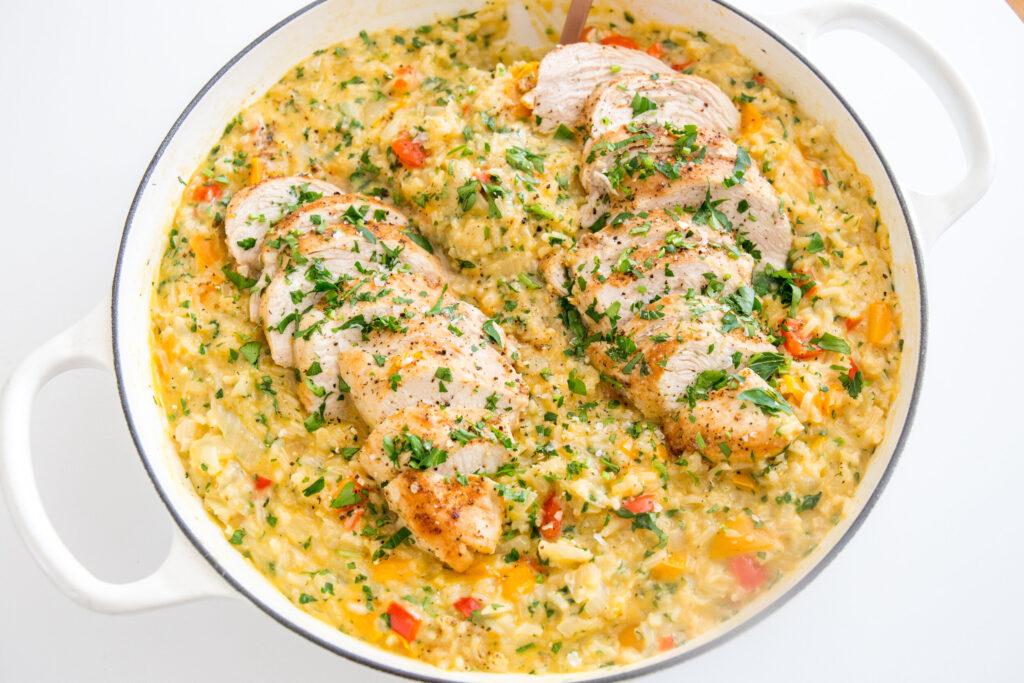 Sliced cooked chicken breast on top of creamy rice with chopped vegetables and herbs in a large white pot.