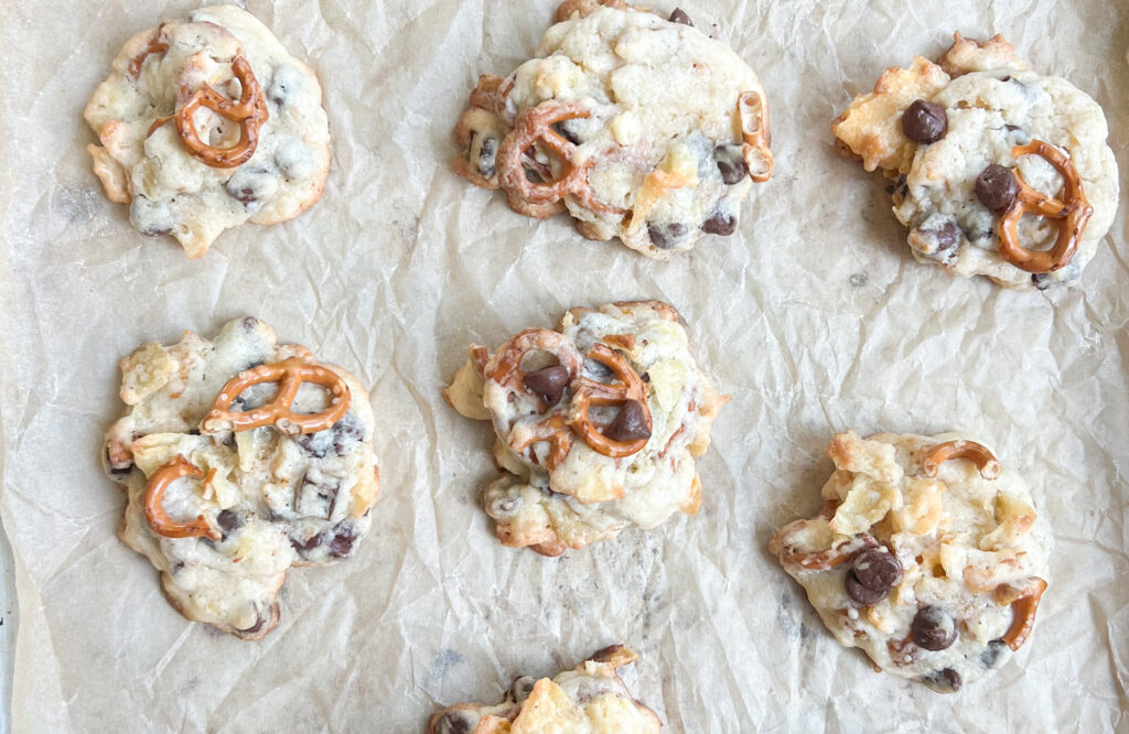 Seven kitchen sink cookies, packed with pretzels, chocolate chips, and potato chips, are arranged on a crumpled sheet of parchment paper.